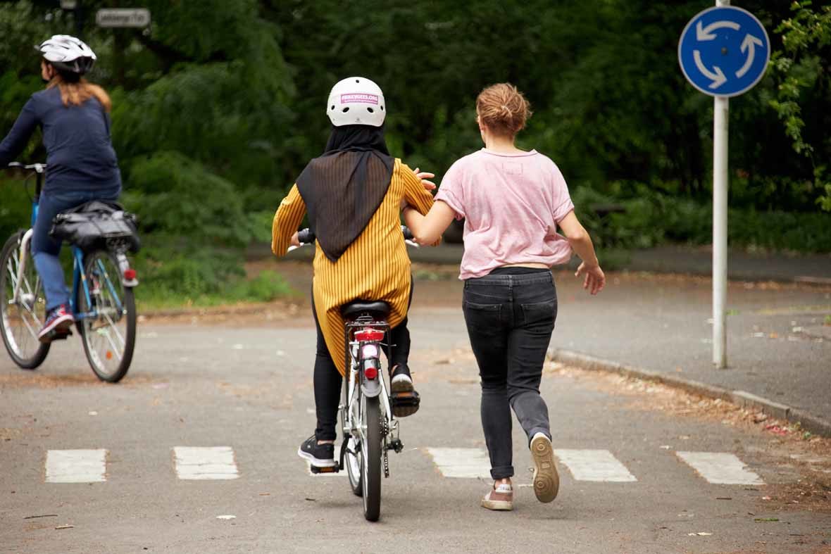 Foto: Eine Frau fährt auf einem Fahrrad, eine andere läuft neben ihr her und stützt sie am Arm
