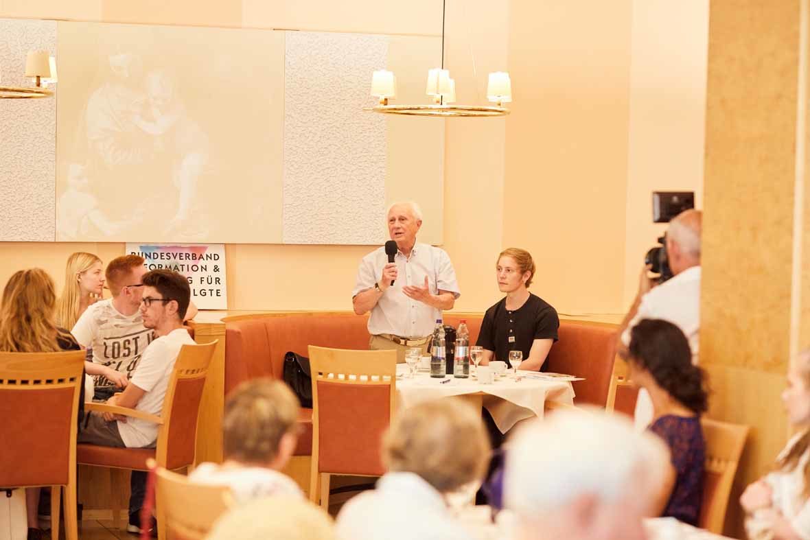Foto: Ein älterer Mann steht in einem großen Raum voller Menschen und hält ein Mikrofon in der Hand und spricht