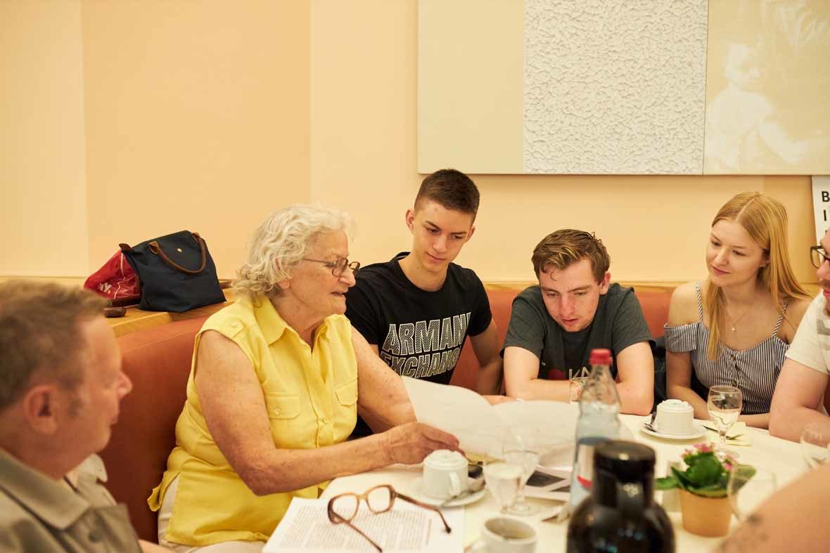 Foto: Eine ältere Frau am Tisch zeigt mehreren Jugendlichen etwas auf einem Papier