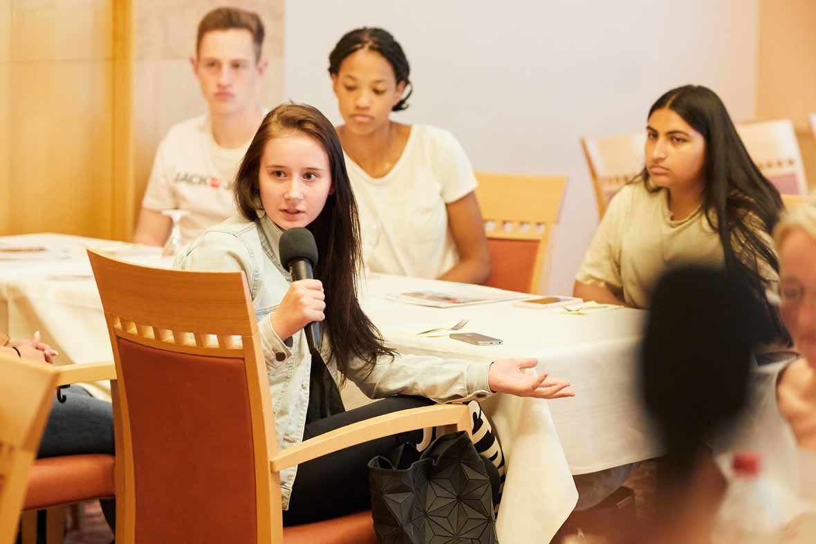 Foto: Eine junge Frau sitzt am Tisch mit einem Mikrofon in der Hand und schaut fragend