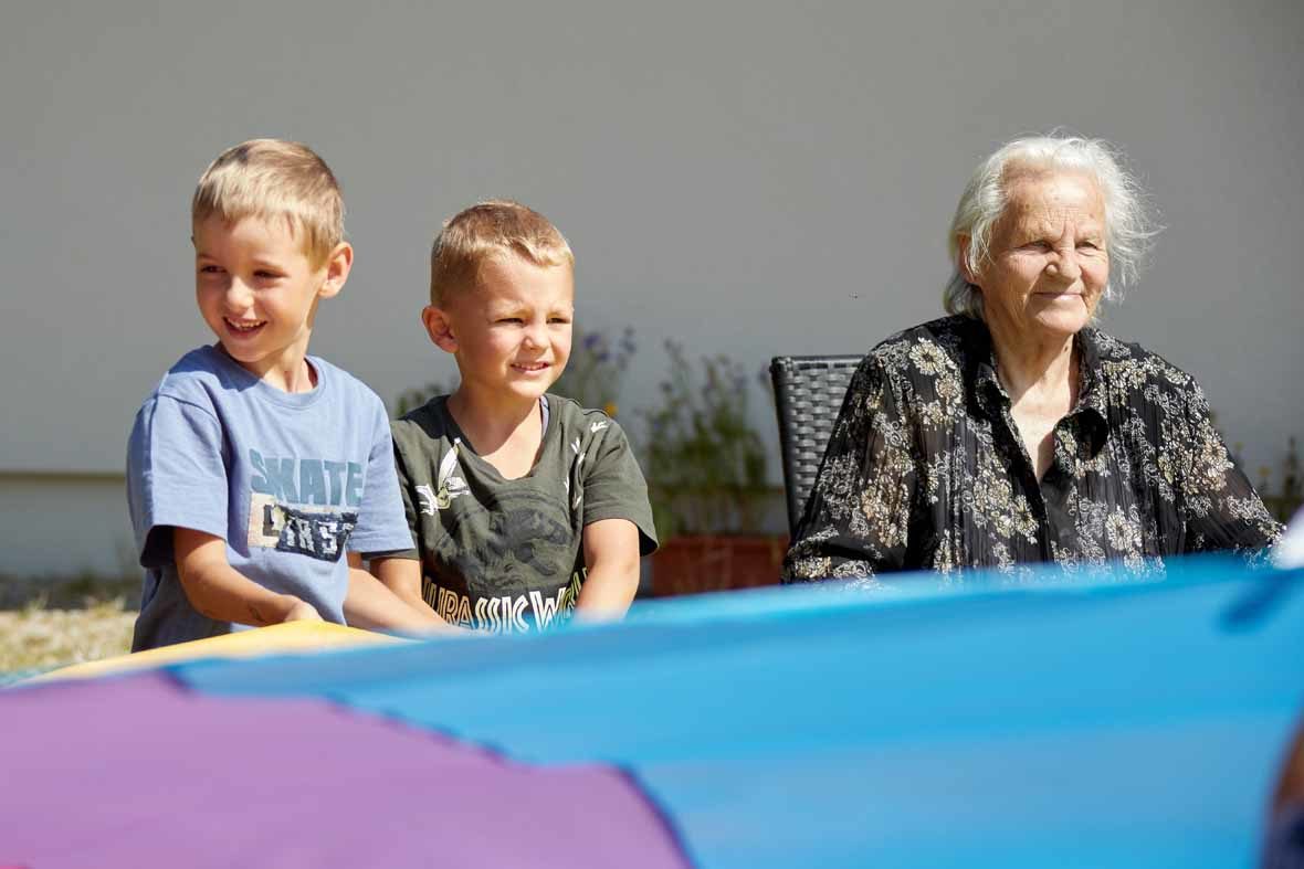 Foto: Zwei Jungen im Kindergartenalter und eine alte Frau halten ein buntes Schwungtuch