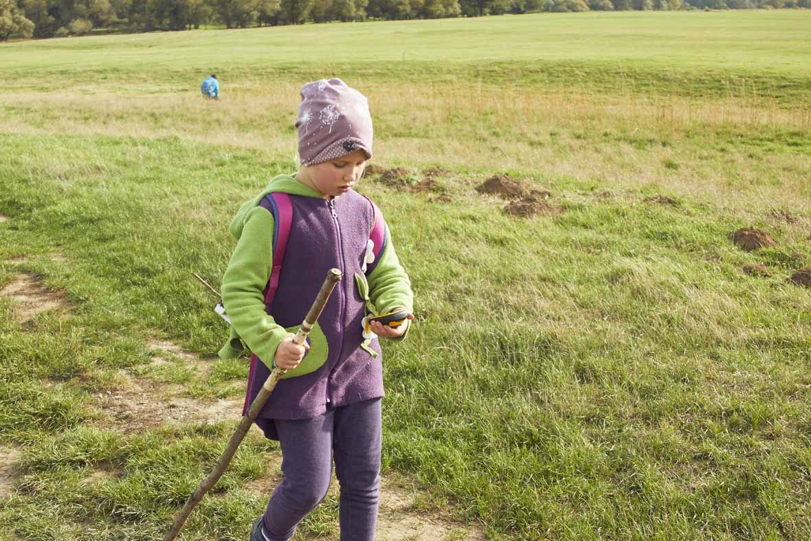Foto: Ein Mädchen mit Wanderstock geht über eine Wiese und schaut auf einen GPS-Tracker in seiner Hand.