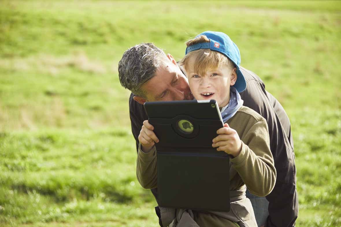 Foto: Ein Junge hält ein Tablet in der Hand, der Vater beugt sich zu ihm.