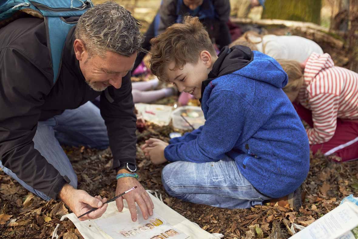 Foto: Ein Vater und sein Sohn bemalen einen Jutebeutel, der auf dem Waldboden liegt.