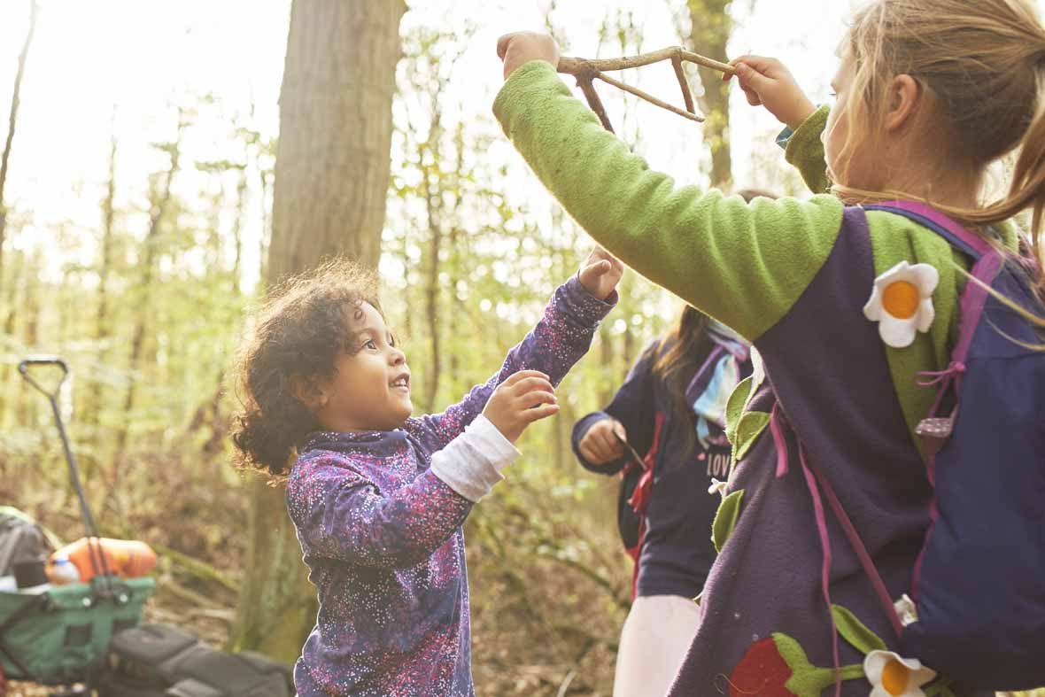 Foto: Drei Mädchen im Wald, eines hält einen Stock hoch, das andere greift danach.