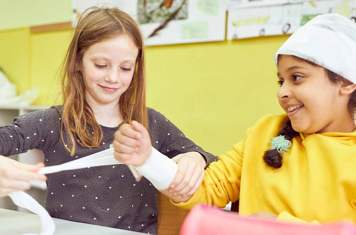 Ein Mädchen legt einen Druckverband am Arm eines anderen Mädchens an.