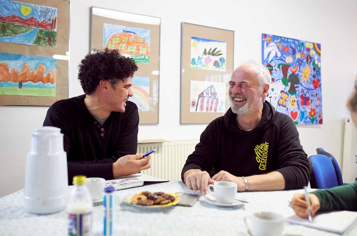Zwei Männer sitzen am Tisch und lachen sich an.