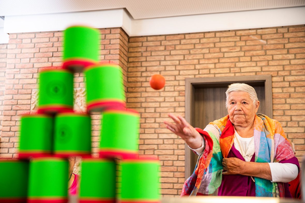Eine ältere Frau wirft mit einem kleinen Ball Schaumstoffdosen um.