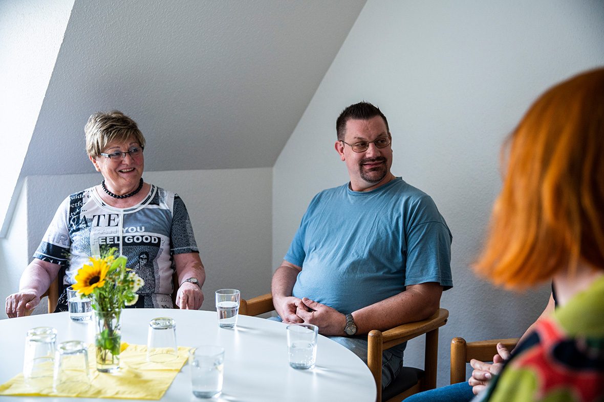 Eine Frau und ein Mann sitzen an einem kleinen Tisch und hören einer dritten Person zu.