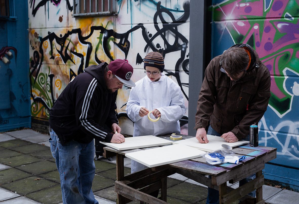 Zwei Graffiti-Künstler kleben mit einem Jungen Holztafeln zum Ansprayen ab.