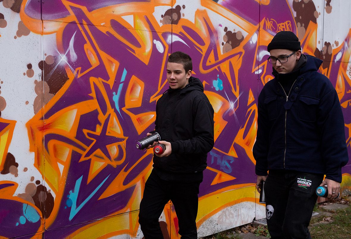 Zwei Jugendliche mit Spraydosen vor einer Wand mit Graffiti.