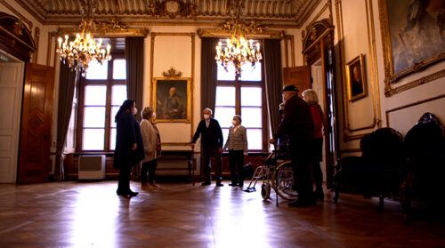 In einem opulent ausgestatteten Museumsraum stehen mehrere ältere Menschen zusammen und unterhalten sich. Die Menschen tragen einen Mund-Nasen-Schutz.
