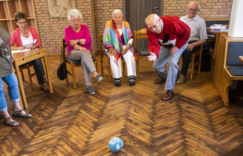 Ein alter Mann wirft einen kleinen Ball wie eine Kegelkugel. Hinter ihm sitzen mehrere Menschen und schauen zu.