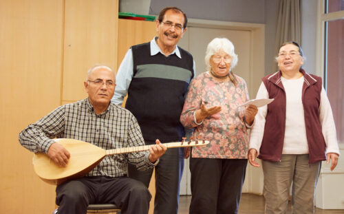 Vier ältere Menschen stehen zusammen und singen. Ein Mann spielt ein Instrument.