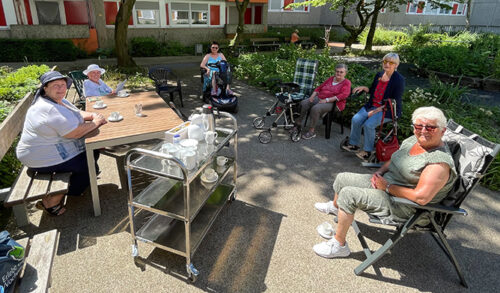Sechs ältere Damen sitzen draußen, trinken Kaffee und eine von ihnen liest etwas vor.