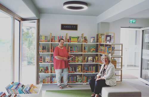 Eine Frau steht vor einem großen Bücherregal, eine andere Frau sitzt davor auf einem Hocker. Eine Frau steht vor einem großen Bücherregal, eine andere Frau sitzt davor auf einem Hocker.